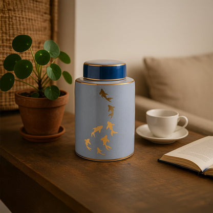 Elegant blue ceramic jar with golden fish design styled on wooden table beside plant and cup for modern decor.