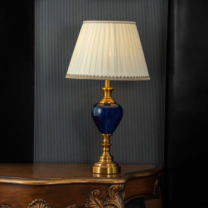 Elegant blue glass table lamp turned on with warm glow and pleated shade displayed on black background decor.