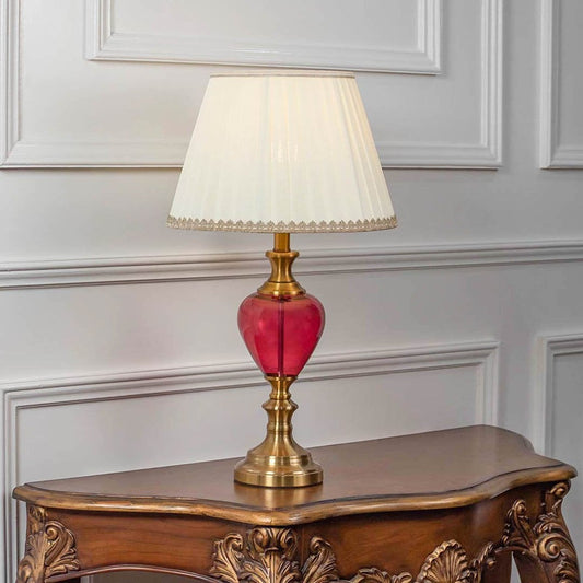 Red glass table lamp turned on with pleated fabric shade and brass detailing displayed on white background.