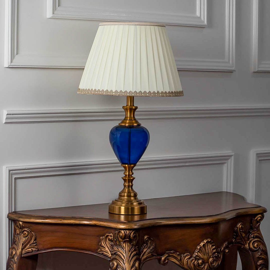 Blue glass table lamp turned off with fabric pleated shade and golden accents displayed on white background.