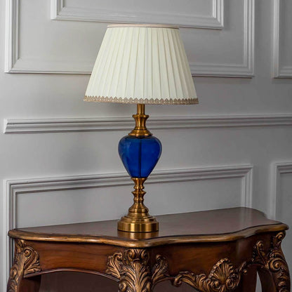 Blue glass table lamp turned off with fabric pleated shade and golden accents displayed on white background.