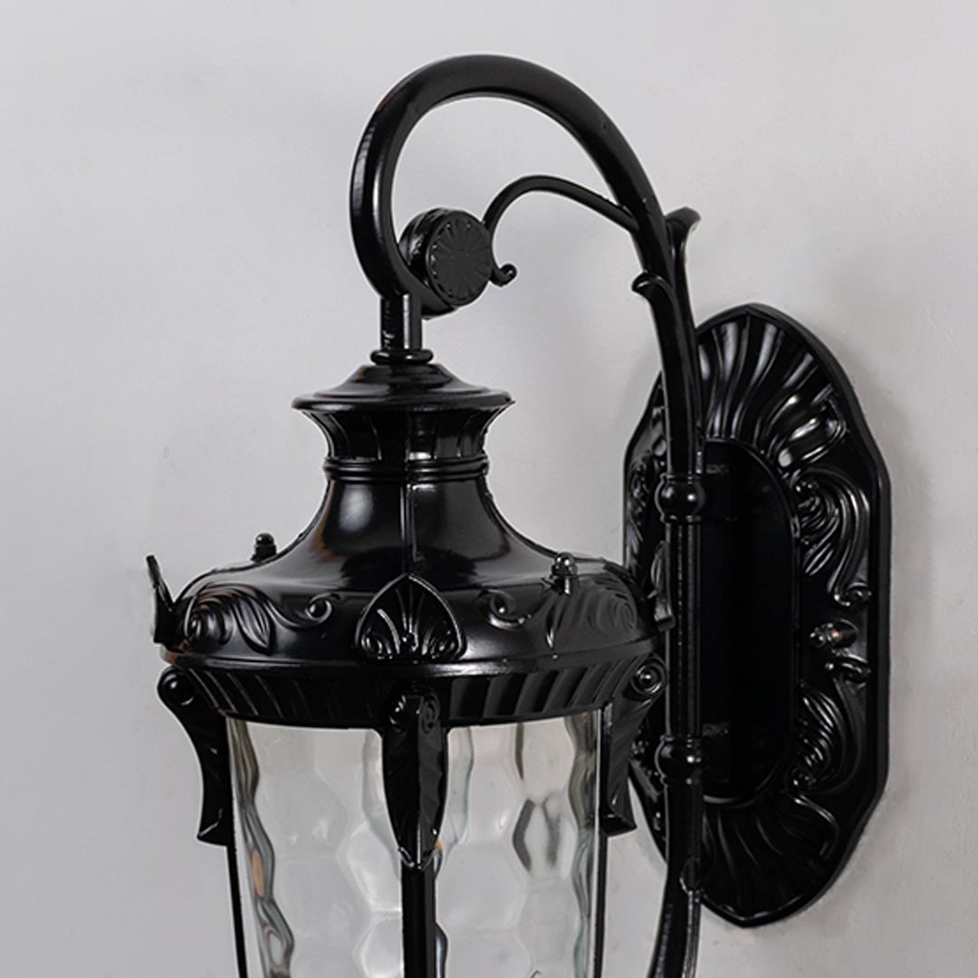 Glossy black metal outdoor wall light closeup image with ornate leaf trim and textured water glass shade for decor.