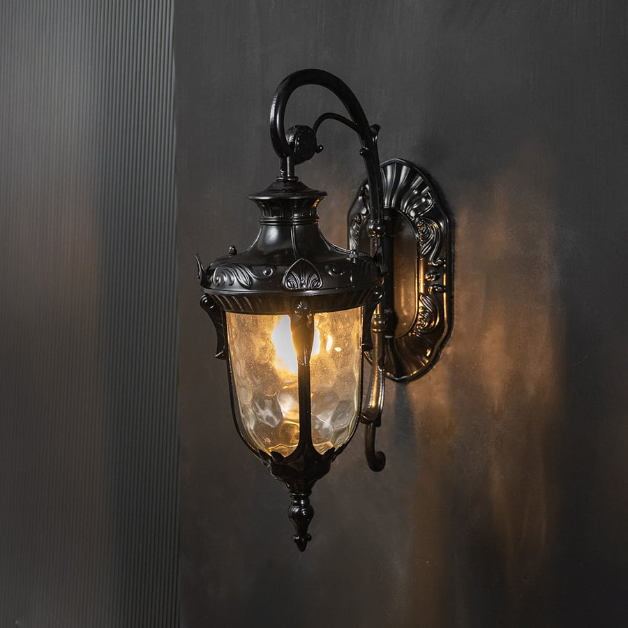 Traditional outdoor wall light with ornate detailing and clear glass glows against a black background.