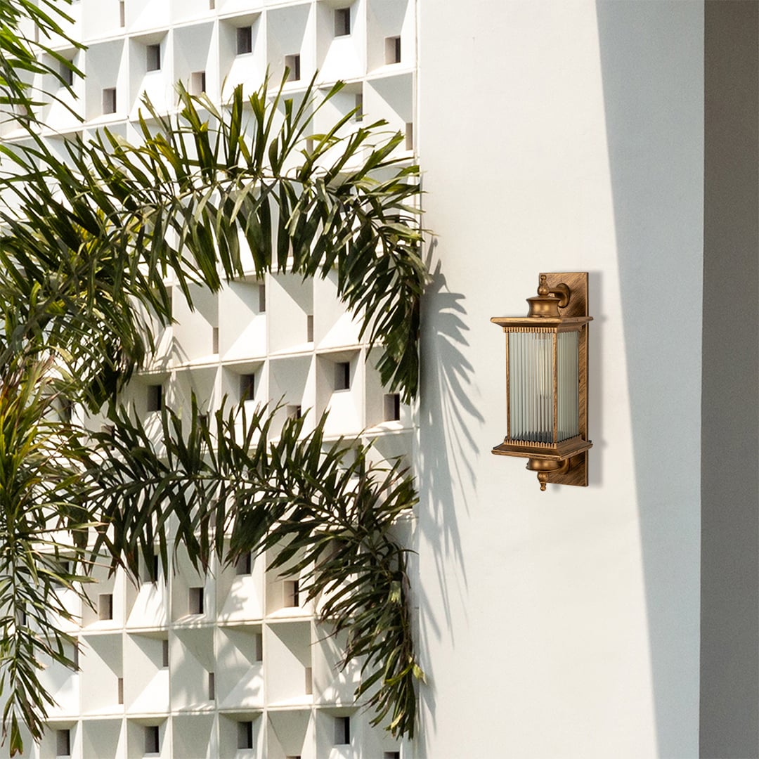 Antique brass metal outdoor wall light lifestyle image with fluted glass shade mounted on white villa wall with palm trees.