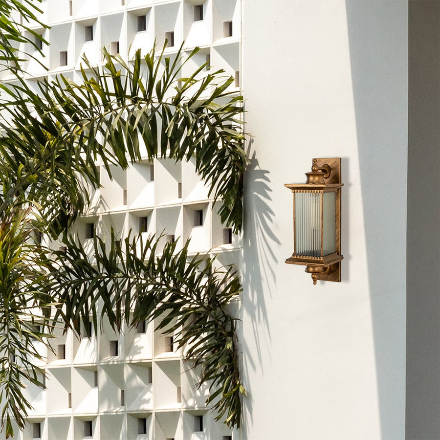 Antique brass metal outdoor wall light lifestyle image with fluted glass shade mounted on white villa wall with palm trees.