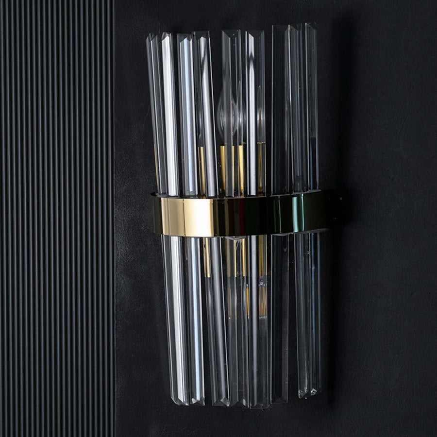 Crystal wall light with the light on against a black background creating a dramatic glow that enhances the reflective glass rods and golden band.