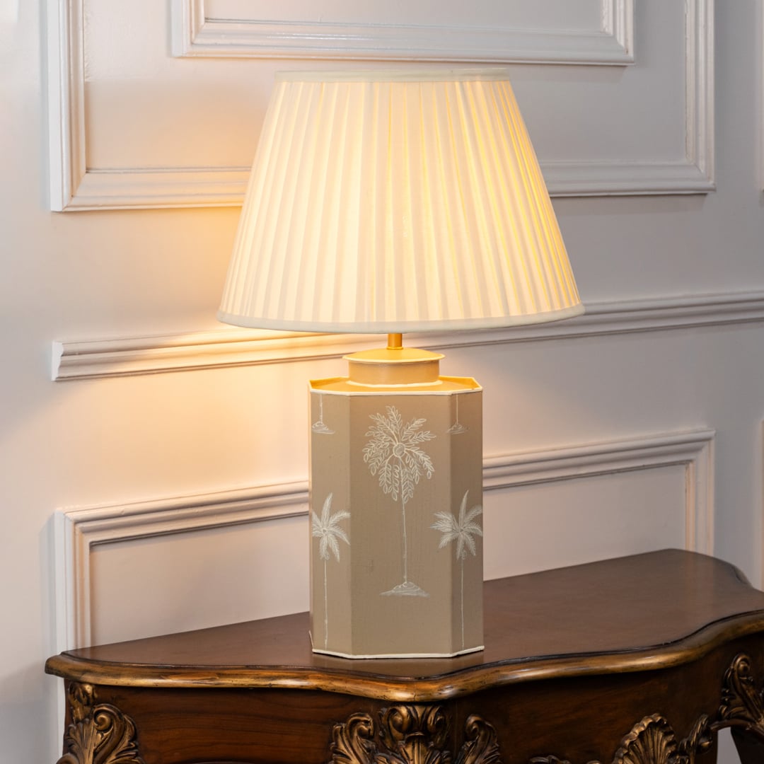 Decorative table lamp with pleated white shade and beige ceramic base featuring subtle palm tree motifs and soft gold accents.