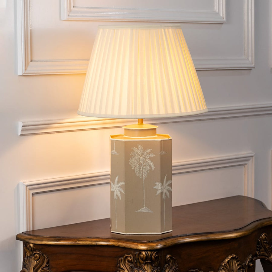 Decorative table lamp with pleated white shade and beige ceramic base featuring subtle palm tree motifs and soft gold accents.