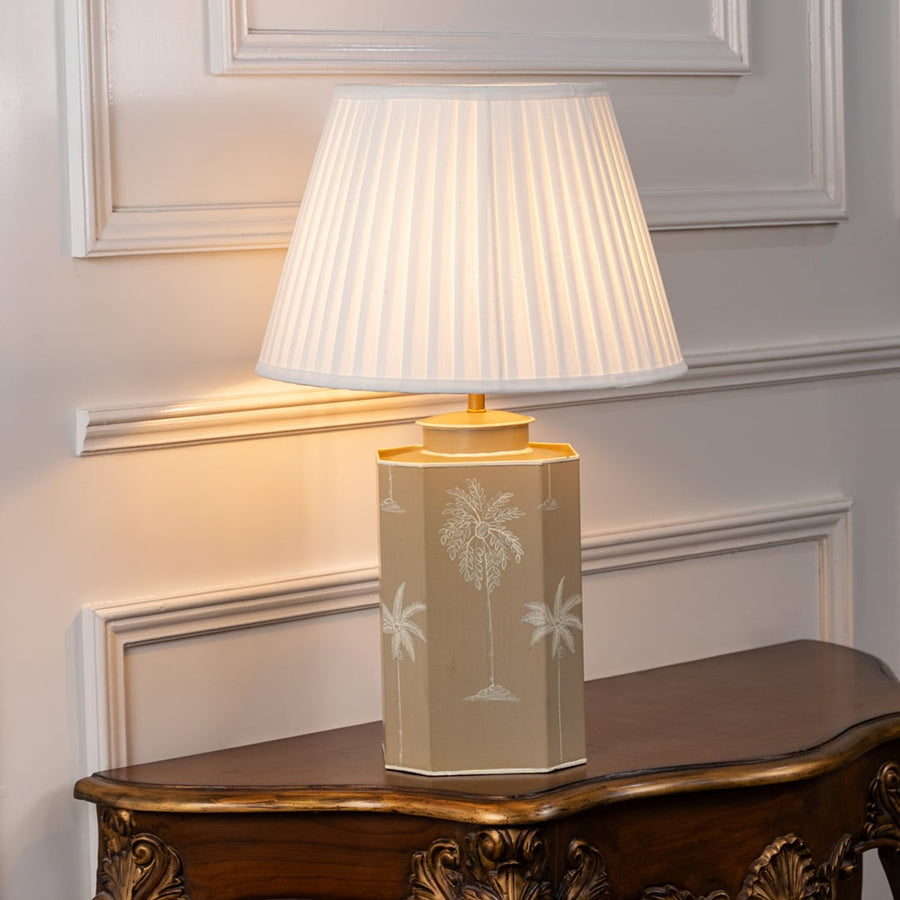 Table lamp with white pleated shade and beige ceramic base featuring subtle palm motif and gold accents styled on wooden table.