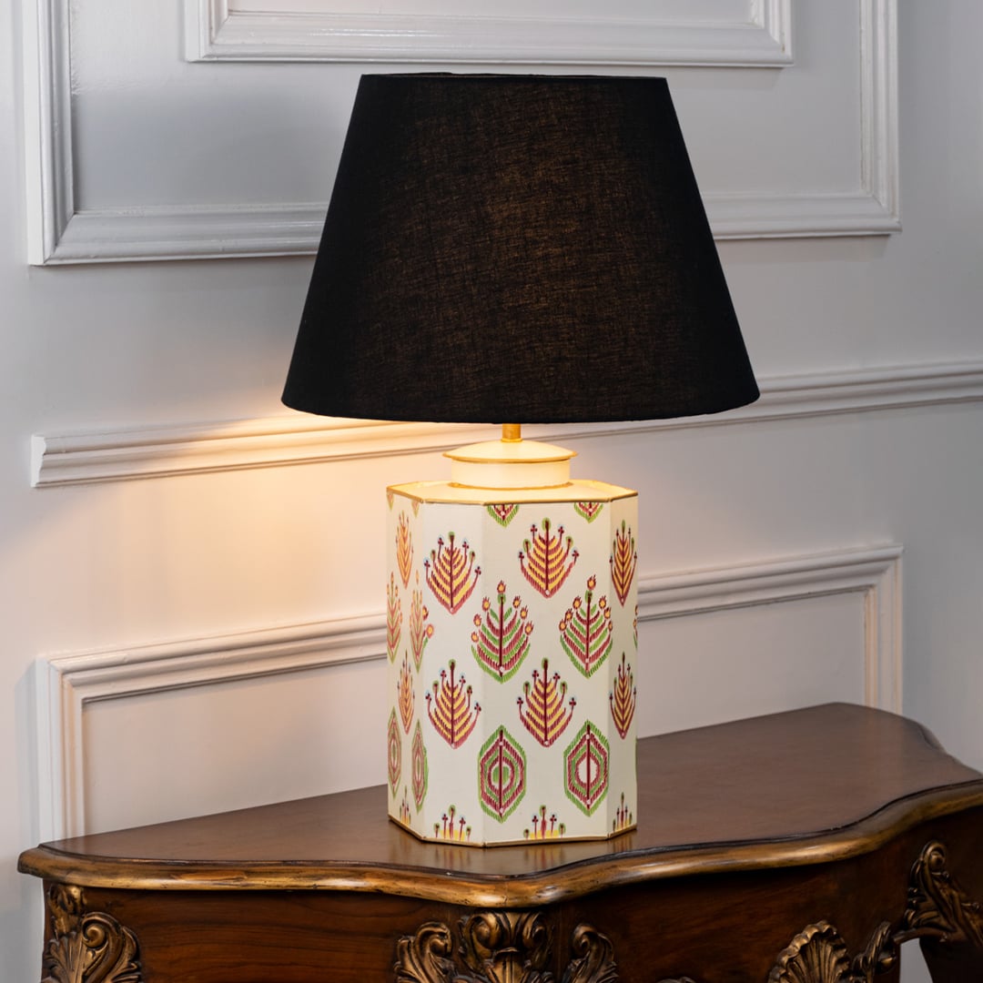 Table lamp with black fabric shade and off white ceramic base decorated with traditional leaf motif patterns styled on a wooden table against a white background.