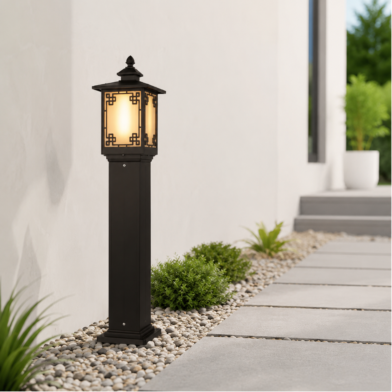 A black outdoor solar pathway light with a warm glowing lantern-style shade, installed on a pebble bed beside a concrete walkway in a modern patio setting.