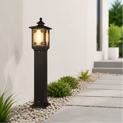A black outdoor solar pathway light with a warm glowing lantern-style shade, installed on a pebble bed beside a concrete walkway in a modern patio setting.