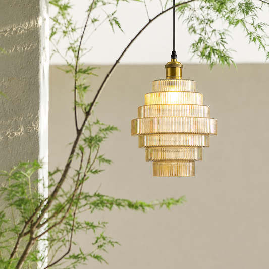 A tiered pendant light with a ribbed glass shade and a brass-toned top cap, hanging from a black cord against a neutral wall with soft-focus green foliage in the foreground.