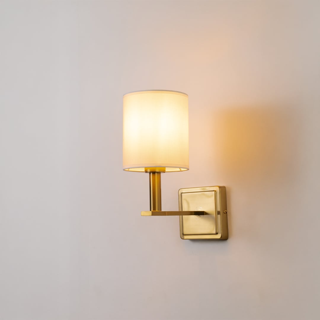 Gold wall sconce with white drum shade glowing softly on white background for modern warm and elegant home lighting decor.