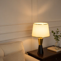 White shade black and gold table lamp on wooden side table with light on beside beige sofa for modern stylish décor.