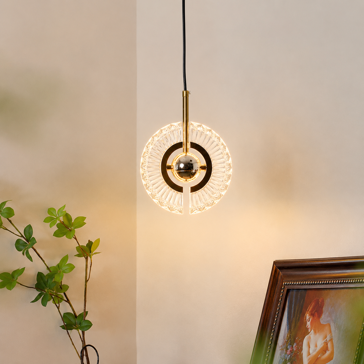 A modern circular pendant light with a gold-finished central rod and clear faceted glass rings, suspended against a warm beige wall. Part of a green plant and a framed picture are visible in the room setting.