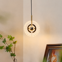 A modern circular pendant light with a gold-finished central rod and clear faceted glass rings, suspended against a warm beige wall. Part of a green plant and a framed picture are visible in the room setting.