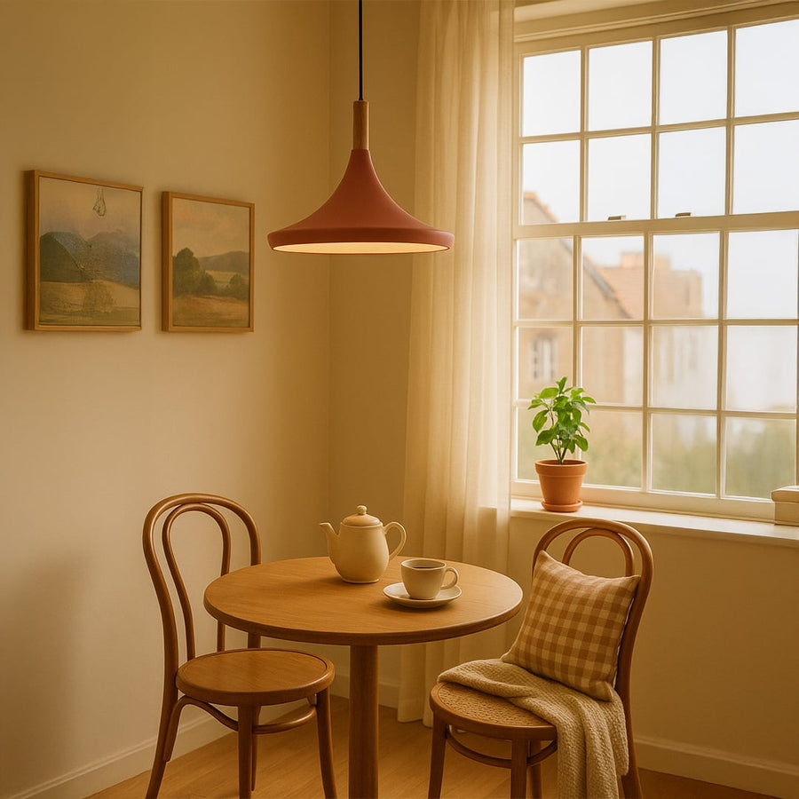 A pink pendant light brightens a cozy dining nook with wooden chairs a tea set and a plant by the window in a warm setting.