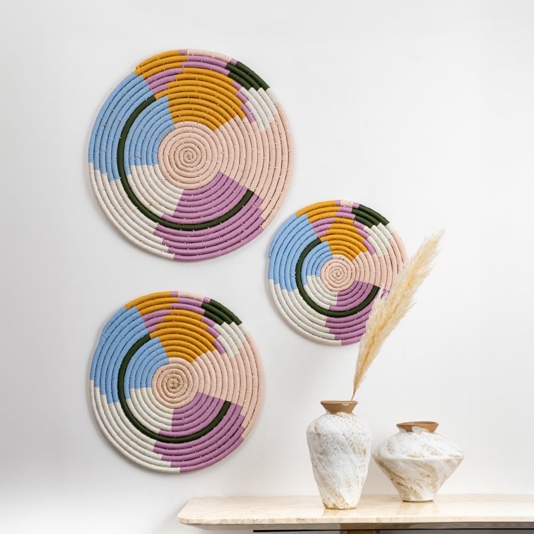 A bright main lifestyle setup showing three multicolor woven wall plates arranged on a white wall above a minimal table with decorative vases creating a warm artistic display.