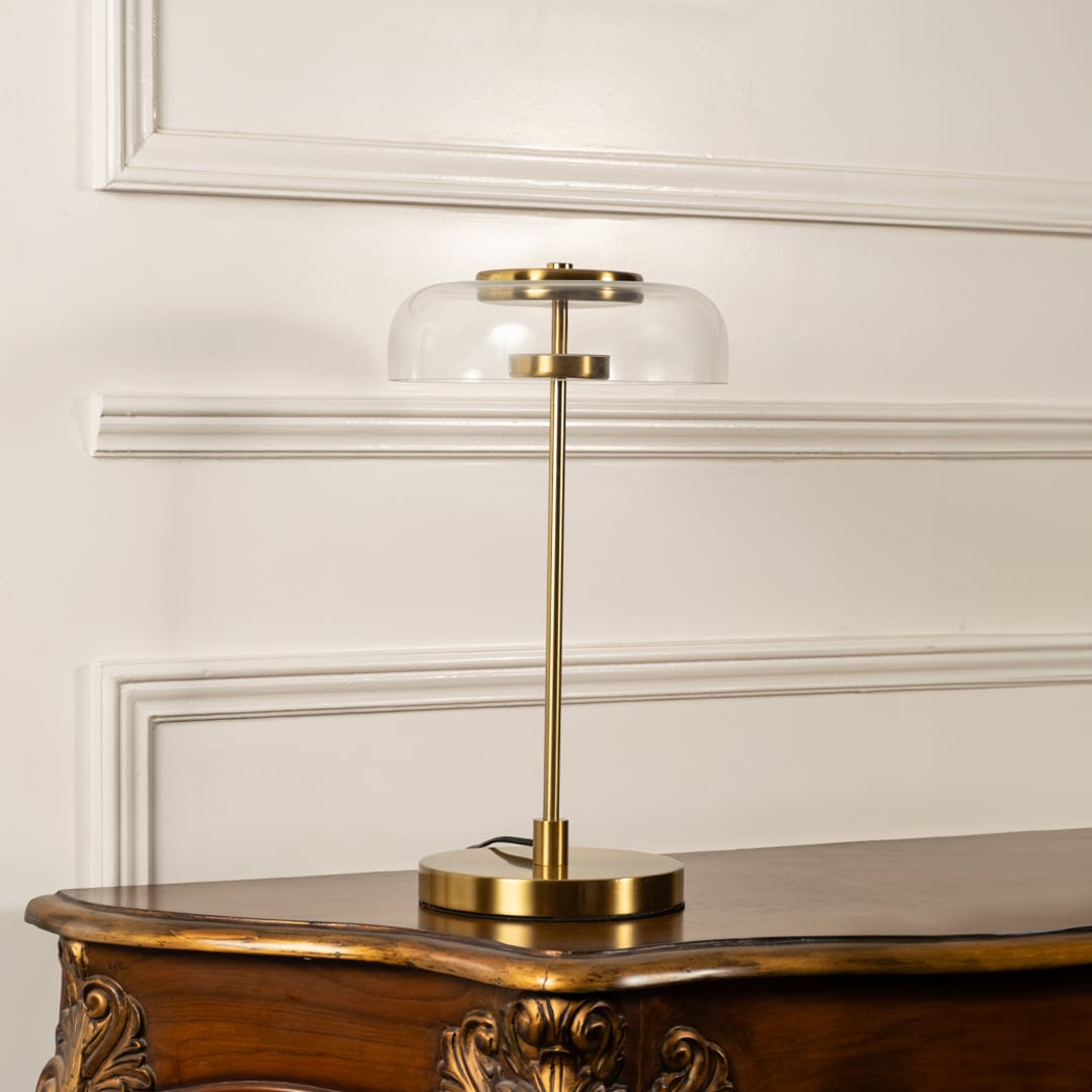 Gold table lamp with illuminated clear glass dome shade and slim metal stem shown on a white background ideal for bedside tables and modern decor.