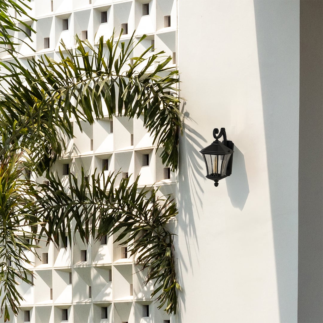 A decorative black outdoor wall lantern with the light off mounted on a bright exterior wall surrounded by lush greenery highlighting its curved arm and elegant lantern frame.