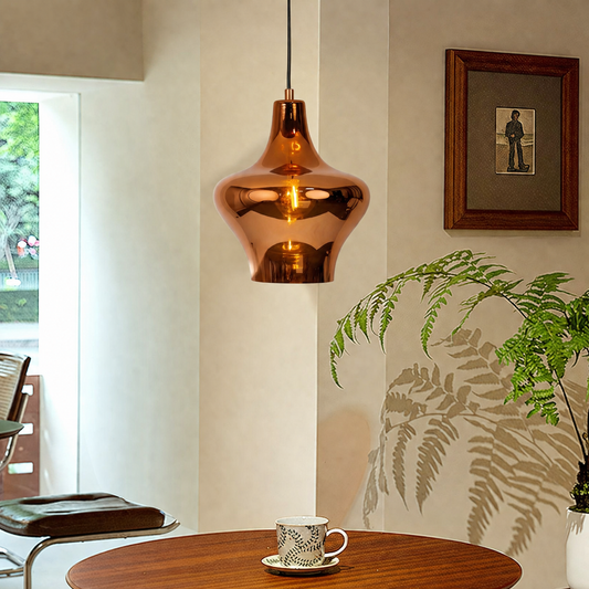 A copper-colored metallic pendant light hangs over a wooden table in a modern interior setting with a coffee cup, a potted fern, and a framed picture on the wall.