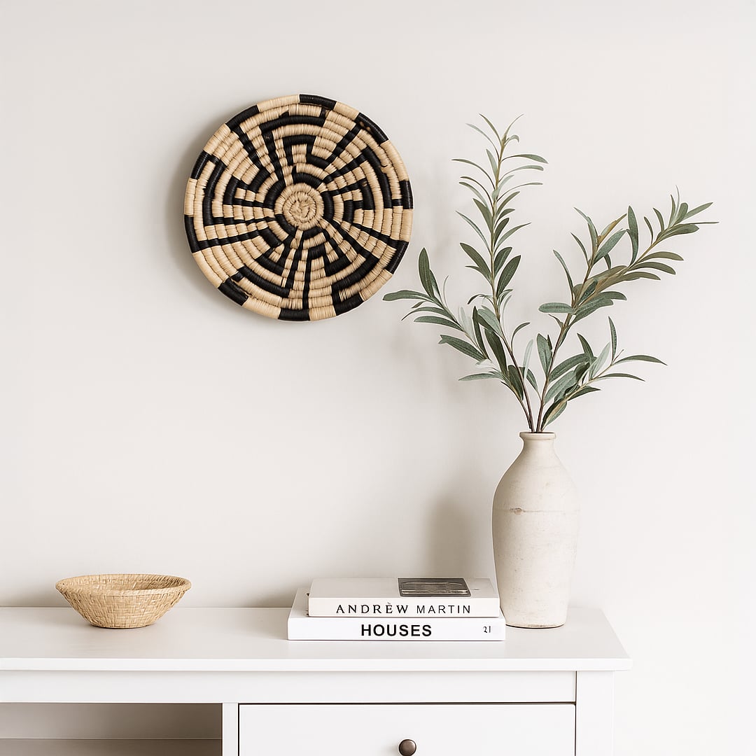 A handwoven natural and black rattan wall decor plate styled above a white console with a ceramic vase books and a woven bowl creating warm minimal home decor vibes.