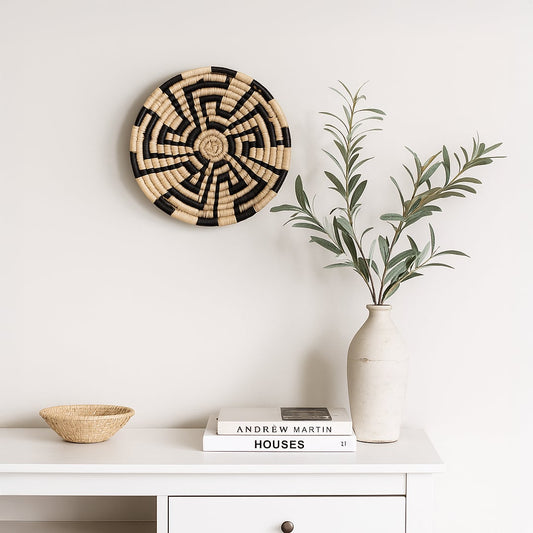 A handwoven natural and black rattan wall decor plate styled above a white console with a ceramic vase books and a woven bowl creating warm minimal home decor vibes.