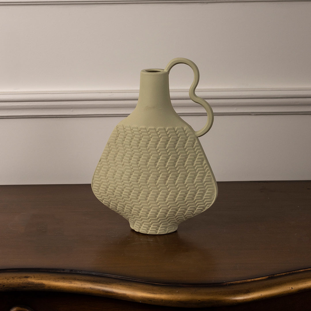 Light yellow ceramic vase with textured pattern and curved handle placed on wooden table against neutral wall background for stylish modern home decor accent.