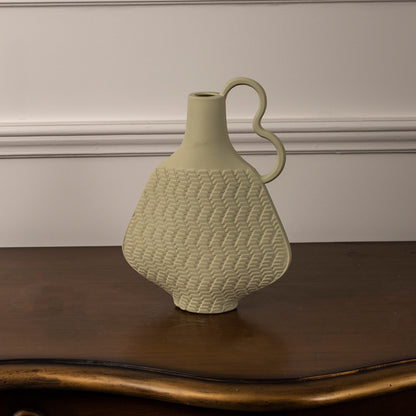 Light yellow ceramic vase with textured pattern and curved handle placed on wooden table against neutral wall background for stylish modern home decor accent.