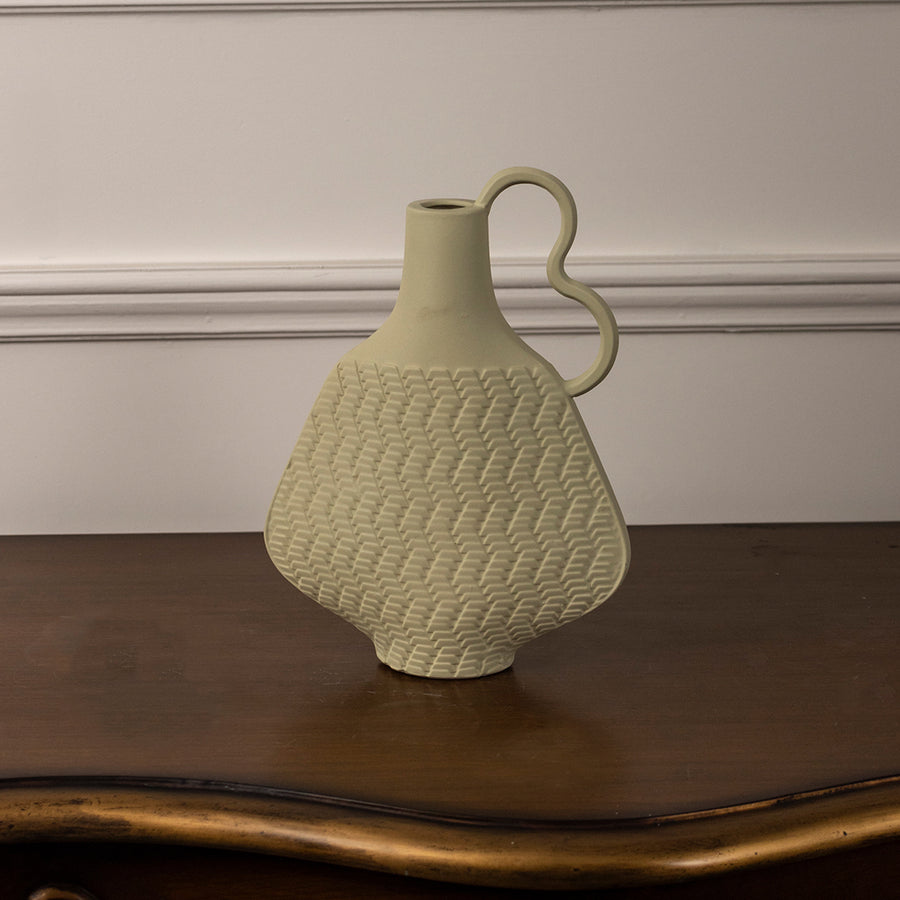 Light yellow ceramic vase with textured pattern and curved handle placed on wooden table against neutral wall background for stylish modern home decor accent.