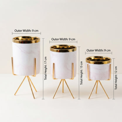 Three white marble-patterned cylindrical planters with gold metal rims and tripod stands, shown in different heights with overlaid dimension labels on a light neutral background.