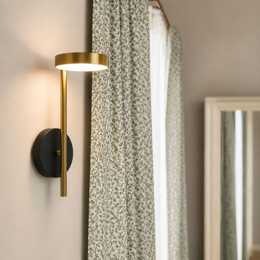 A modern wall sconce light fixture with a matte black circular wall mount and a brushed gold vertical bar and shade, shown lit in a bedroom setting next to patterned curtains.