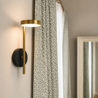 A modern wall sconce light fixture with a matte black circular wall mount and a brushed gold vertical bar and shade, shown lit in a bedroom setting next to patterned curtains.