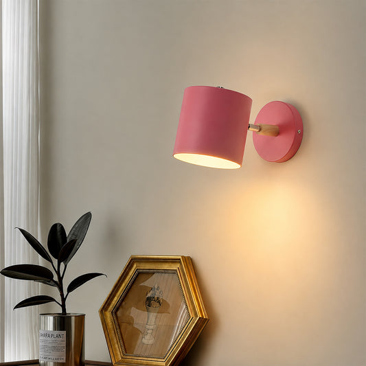 A pink wall-mounted lamp with a cylindrical shade and a gold-finished socket, shown lit on a beige wall above a console table with a plant and framed picture.