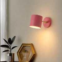 A pink wall-mounted lamp with a cylindrical shade and a gold-finished socket, shown lit on a beige wall above a console table with a plant and framed picture.