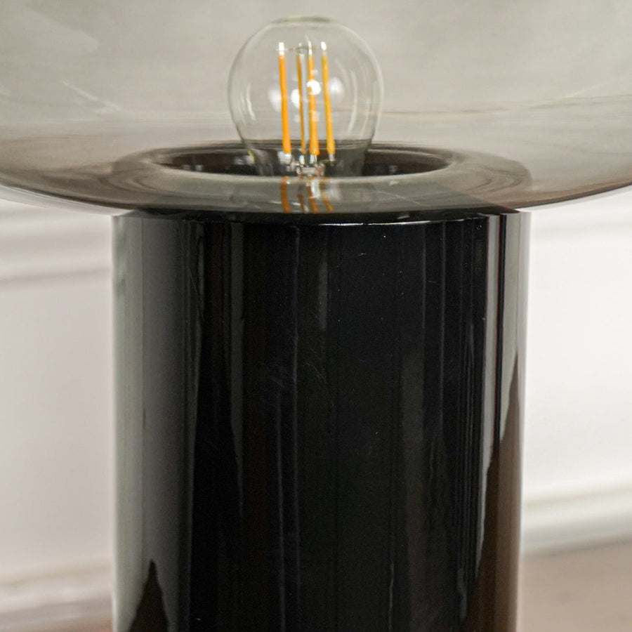 Close up black metal base table lamp with exposed filament bulb and glass shade positioned on wooden surface against neutral wall background.