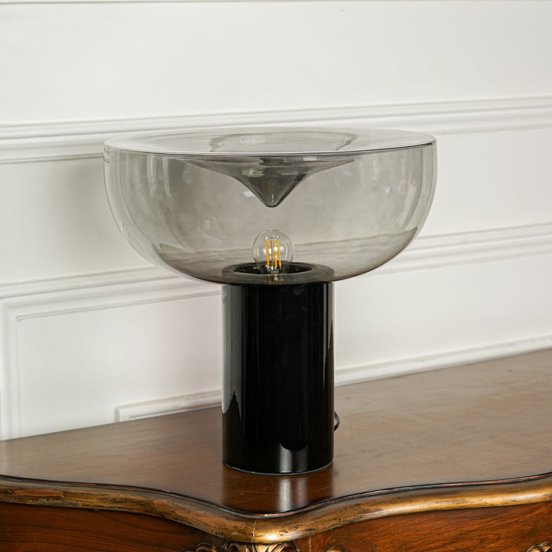 Modern smoked glass table lamp with black metal base placed on wooden surface against textured white wall background ideal for luxury home decor.