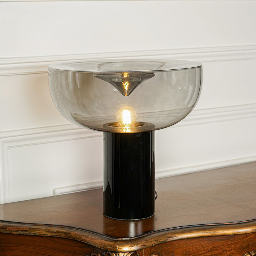 Elegant tinted glass table lamp with gold metal stem and black base positioned on wooden surface against minimal wall background for ambient lighting.
