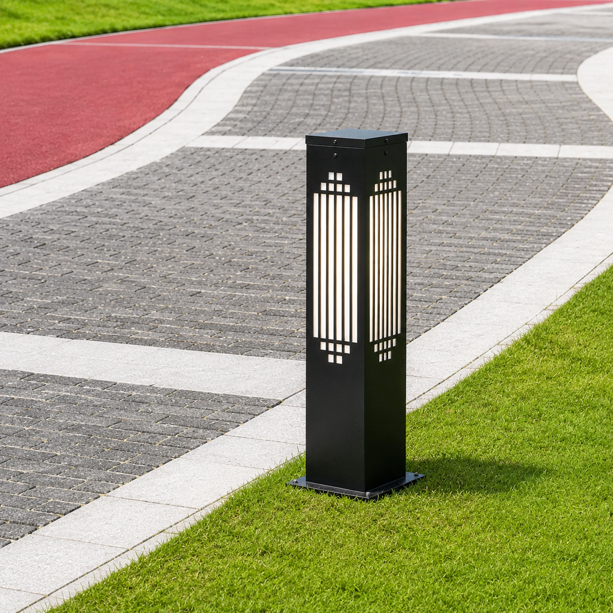 A modern black outdoor bollard post light with vertical slits, standing on a green grass verge next to a paved pathway in a park.