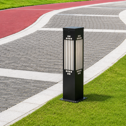 A modern black outdoor bollard post light with vertical slits, standing on a green grass verge next to a paved pathway in a park.