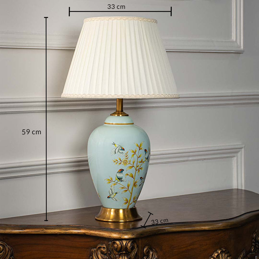 Ceramic table lamp 59 cm height 33 cm width with pleated shade blue base floral bird pattern and gold finish light off white background.