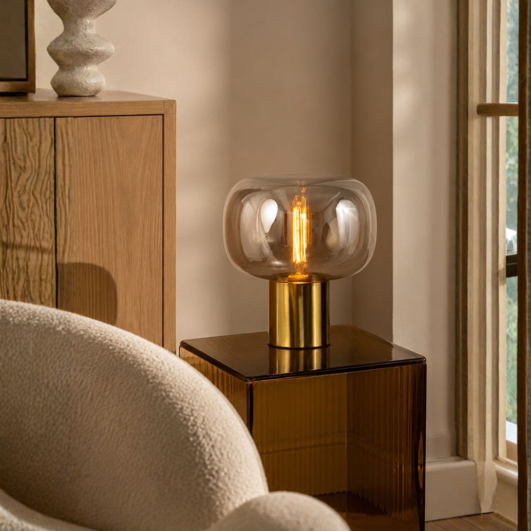 A modern table lamp with a clear rounded glass shade and a brushed gold cylindrical base, lit with a warm Edison-style bulb, placed on a dark side table in a softly lit interior room.