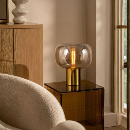 A modern table lamp with a clear rounded glass shade and a brushed gold cylindrical base, lit with a warm Edison-style bulb, placed on a dark side table in a softly lit interior room.