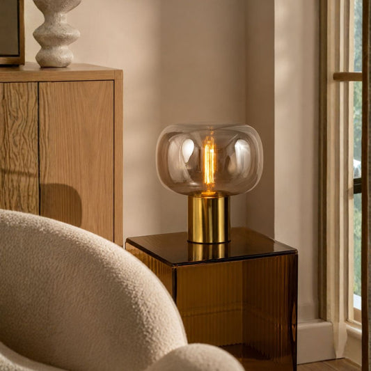 A modern table lamp with a clear rounded glass shade and a brushed gold cylindrical base, lit with a warm Edison-style bulb, placed on a dark side table in a softly lit interior room.