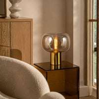A modern table lamp with a clear rounded glass shade and a brushed gold cylindrical base, lit with a warm Edison-style bulb, placed on a dark side table in a softly lit interior room.