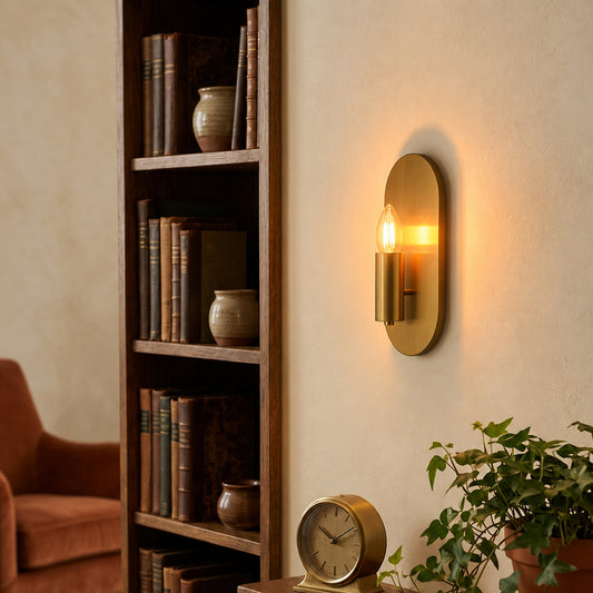 A modern wall-mounted sconce with a warm brass finish and a glowing light bulb, mounted on a beige wall in a cozy room setting next to a wooden bookshelf.