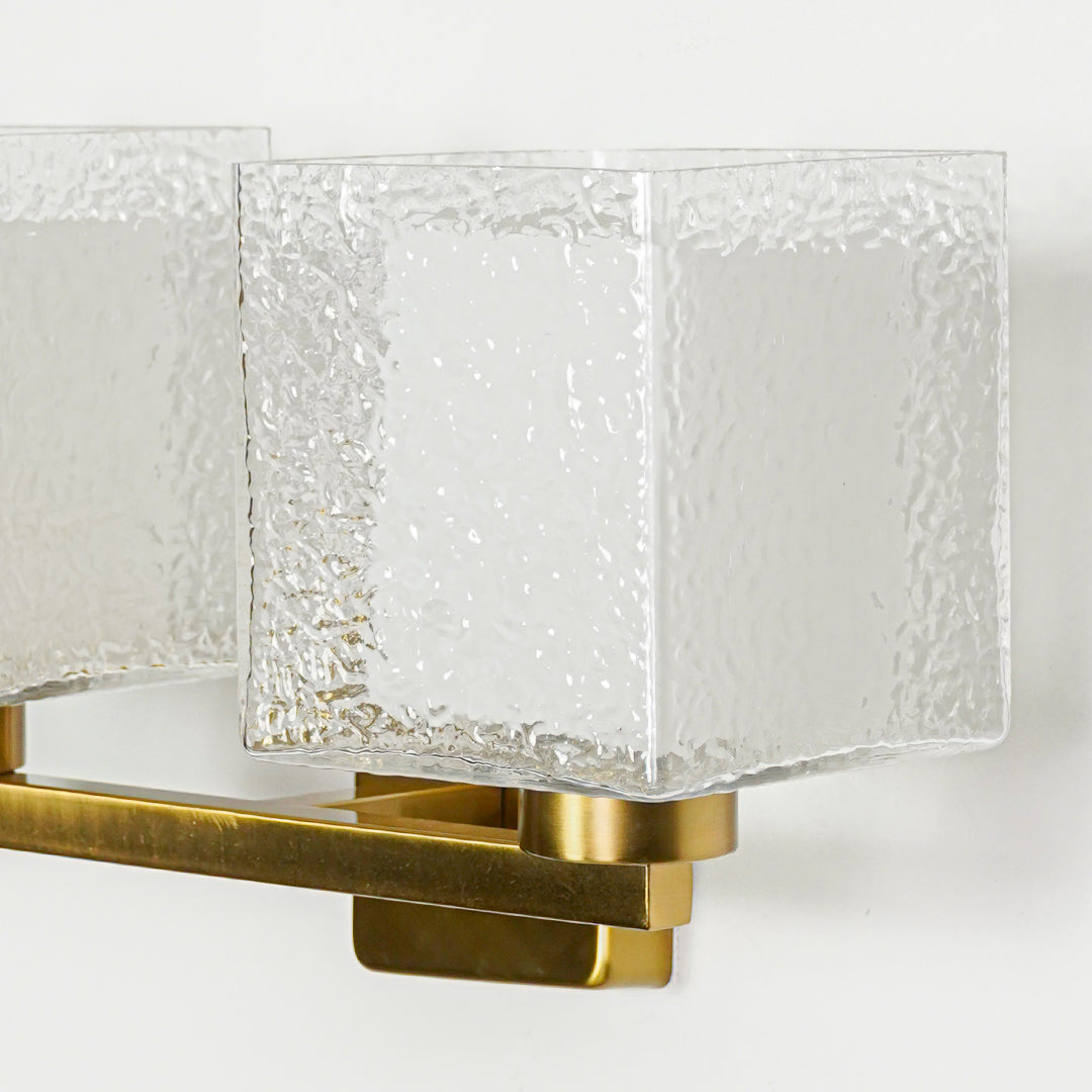 Close view of gold wall light with textured glass cube shade showcasing premium modern lighting design on neutral background.