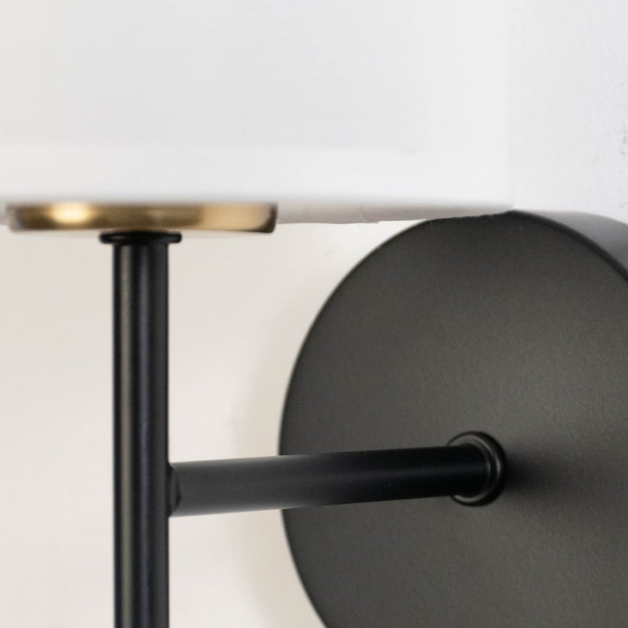 Detailed closeup of white fabric drum shade with brass rim and black metal rod hardware on modern minimalist wall sconce.