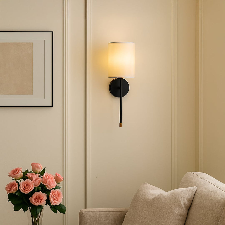 Elegant white fabric cylinder shade wall sconce with black metal arm and brass accents mounted on interior wall lighting.